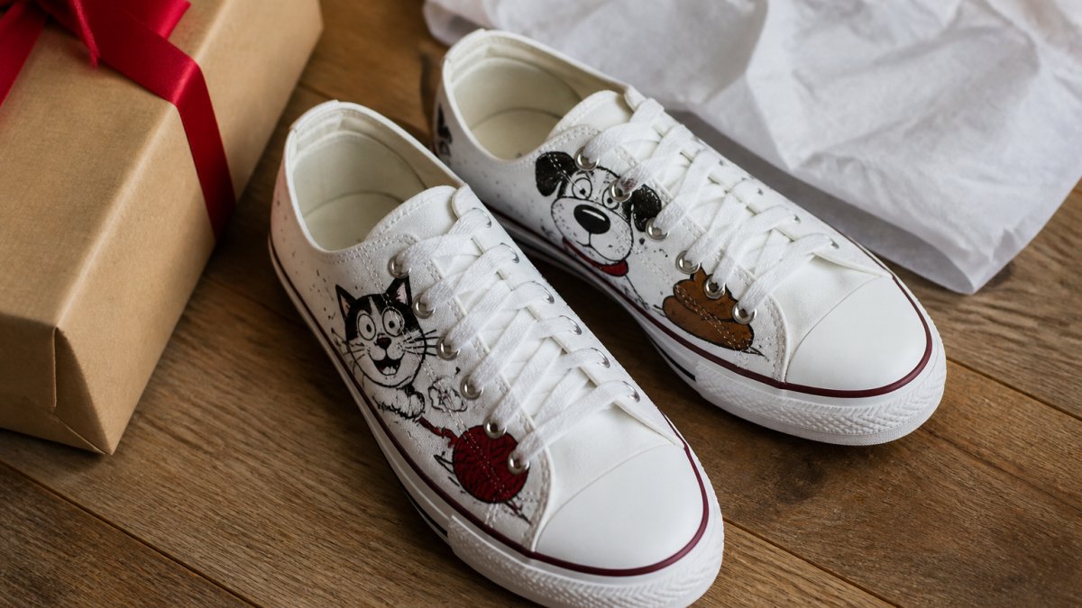A pair of white canvas sneakers with bold cartoon and humorous printed graphics resting on a wooden floor next to a gift box and tissue paper