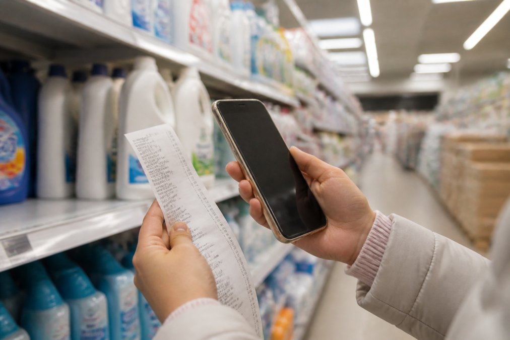 Close-up of hands holding a receipt and comparing prices on a smartphone while shopping for household supplies in a store aisle, with fluorescent lighting overhead