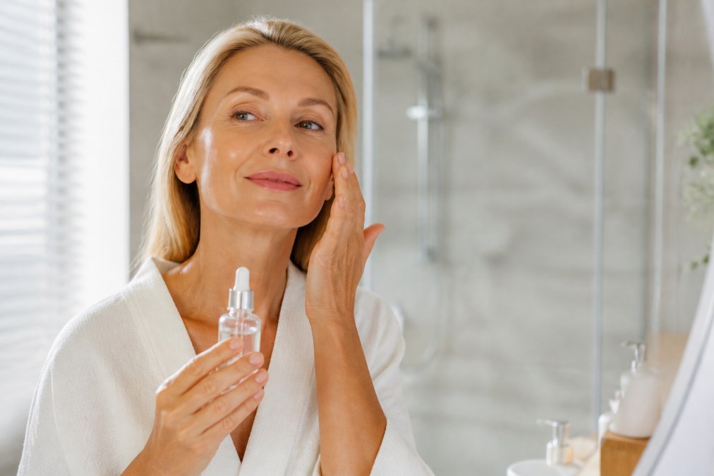 Woman in her 50s applying facial serum to her face in a bright, naturally-lit bathroom, holding a bottle of hydrating serum with visible satisfaction
