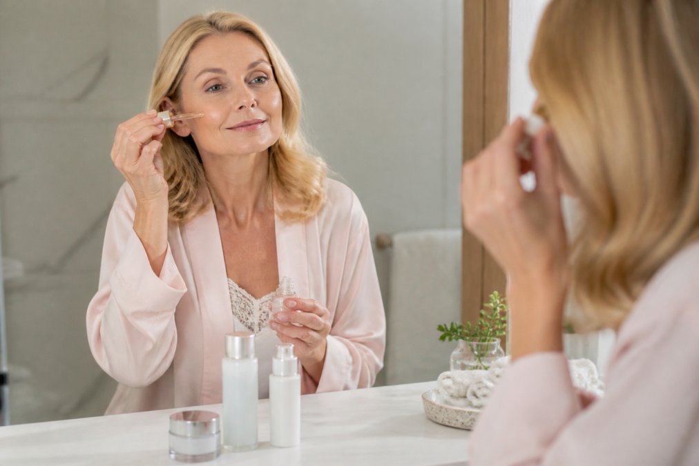 Woman in her 50s with sensitive aging skin gently applying a serum in a softly lit bathroom.