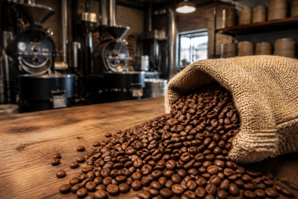 Overhead shot of a burlap coffee bag with roasted beans spilling onto a wooden countertop, a small roastery visible in the background with warm lighting