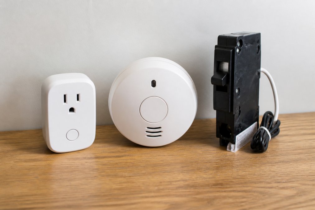 A labeled comparison graphic showing a smart plug, a hardwired smoke detector, and a smart circuit breaker side by side, with icons indicating power source and maintenance needs