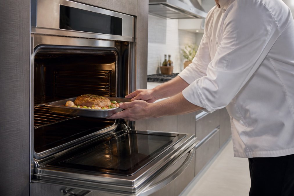 Chef's hands placing a tray into a modern wall oven with a digital touchscreen displaying cooking presets, bright renovated kitchen with stainless steel finishes