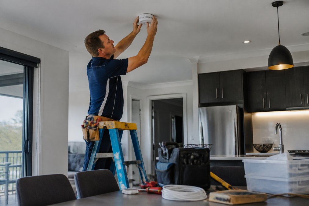 A qualified electrician in a branded work uniform installing a smart smoke alarm on a ceiling inside a modern home, with a step ladder and tools visible nearby and warm interior lighting