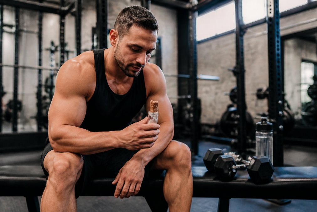 Muscular athlete in gym holding high-protein snack bar with dumbbells and water bottle visible on workout bench, natural gym lighting