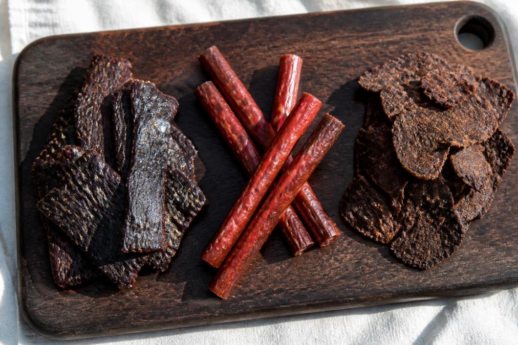 Flat lay of grass-fed beef jerky, beef sticks, and crispy beef crisps arranged on a dark wooden cutting board under bright natural light, with clean white linen background