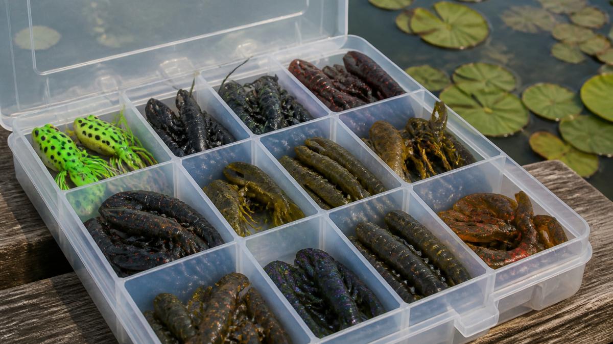 Close-up of a fishing tackle box with multiple colorful soft plastic lures including frogs, craws, and creature baits arranged on a wooden dock with water and lily pads visible in the background