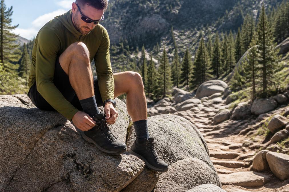 Hiker sitting on a large granite boulder lacing up hiking boots over performance socks, with a steep rocky trail and pine forest visible in the background, warm late-morning light