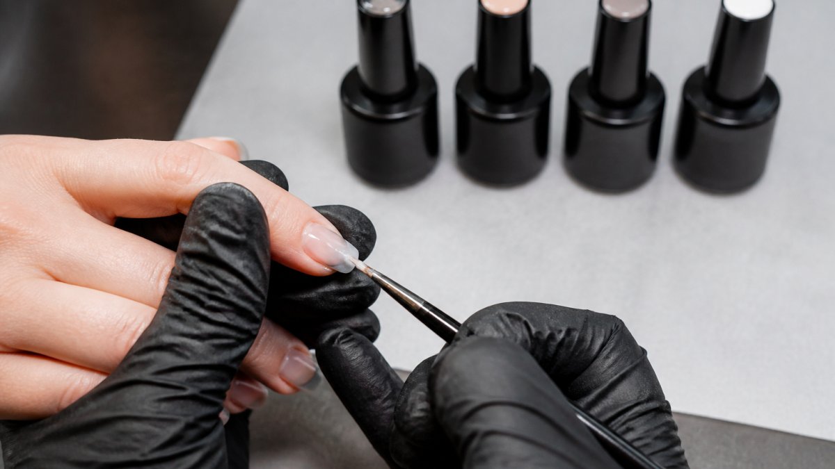 Nail technician applying milky white rubber base gel with a fine liner brush onto a client's natural fingernail