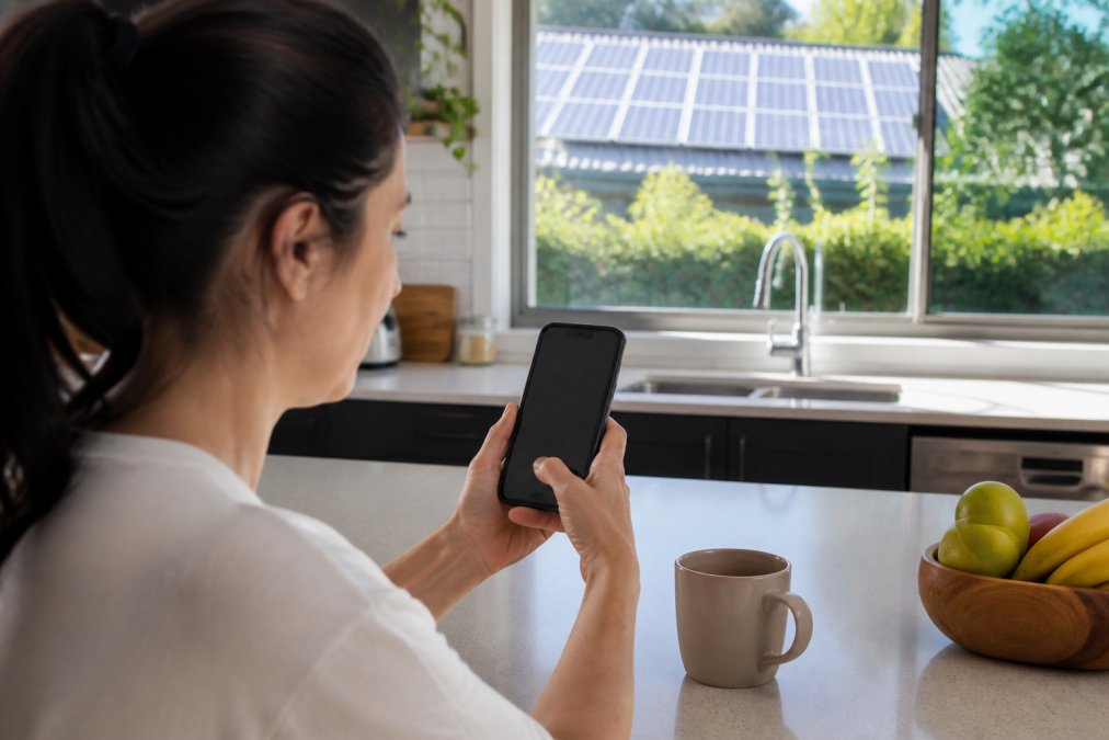 Homeowner viewing solar battery monitoring dashboard on a smartphone in a bright kitchen with solar panels visible through the window, natural sunlight illuminating the screen