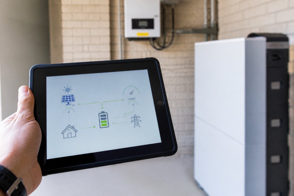 Close-up of a tablet displaying real-time energy flow visualisation with power meter and battery storage unit visible in the background, showing live generation and consumption data