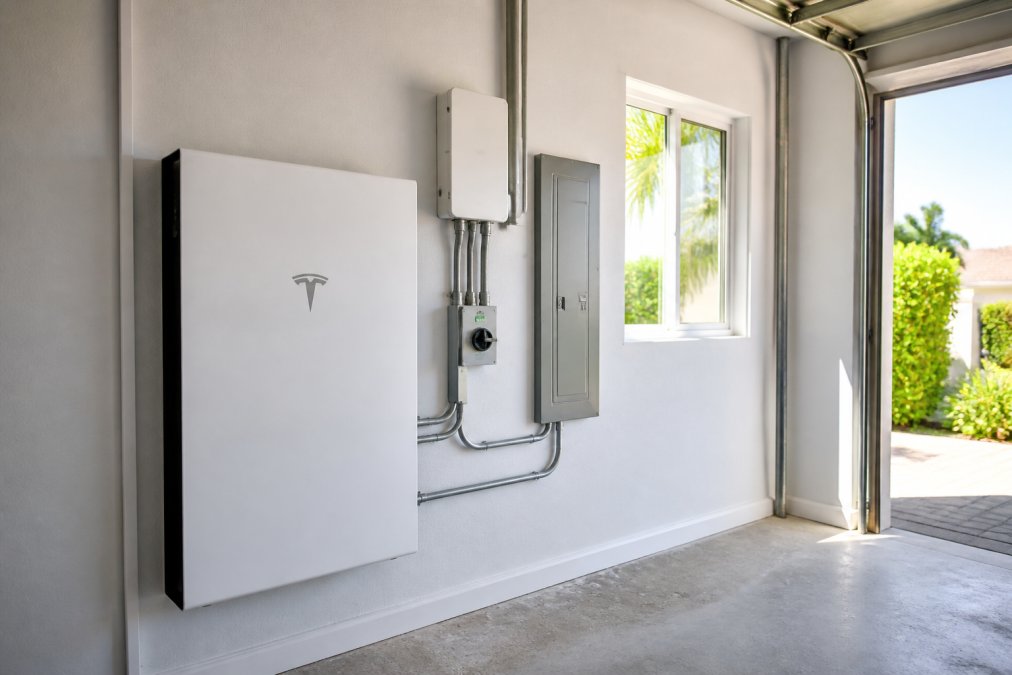Modern residential garage interior showing a sleek white Tesla Powerwall 3 battery unit mounted on the wall beside home electrical infrastructure and circuit breaker panel, natural daylight streaming through a side window