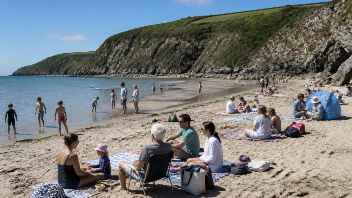 Large multi-generational group enjoying a sunny day on a sandy South Devon beach, with families spread across the sand, children playing in shallow water, and adults relaxing on beach towels, dramatic coastal cliffs in the background