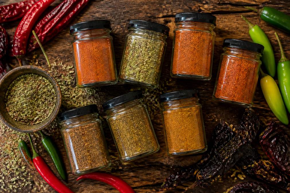Overhead flat-lay of multiple colorful spice jars with handcrafted labels arranged on a rustic wooden surface with fresh red and green chiles and dried Mexican oregano scattered around