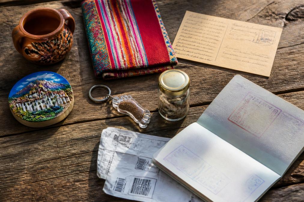 A flat-lay arrangement of colorful travel souvenirs on a weathered wooden surface: small ceramic pottery, a folded textile, a handwritten postcard, a painted fridge magnet, a soil-filled keychain, and a small glass jar, arranged alongside an open passport and a crumpled boarding pass under warm natural light