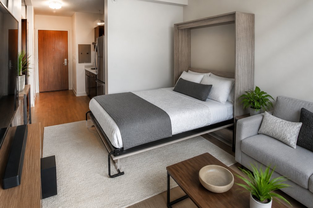 Compact studio apartment interior with a Savona wall bed in the open position showing a queen-sized bed extended horizontally from the wall, with a small living room sofa and entertainment console visible below, demonstrating the dramatic floor space efficiency