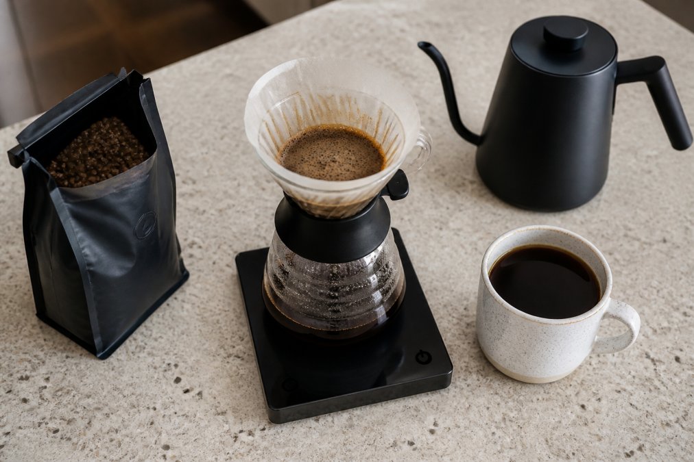 Overhead shot of a kitchen counter with a pour-over dripper, gooseneck kettle, digital scale, and a freshly brewed cup of coffee beside a bag of whole beans