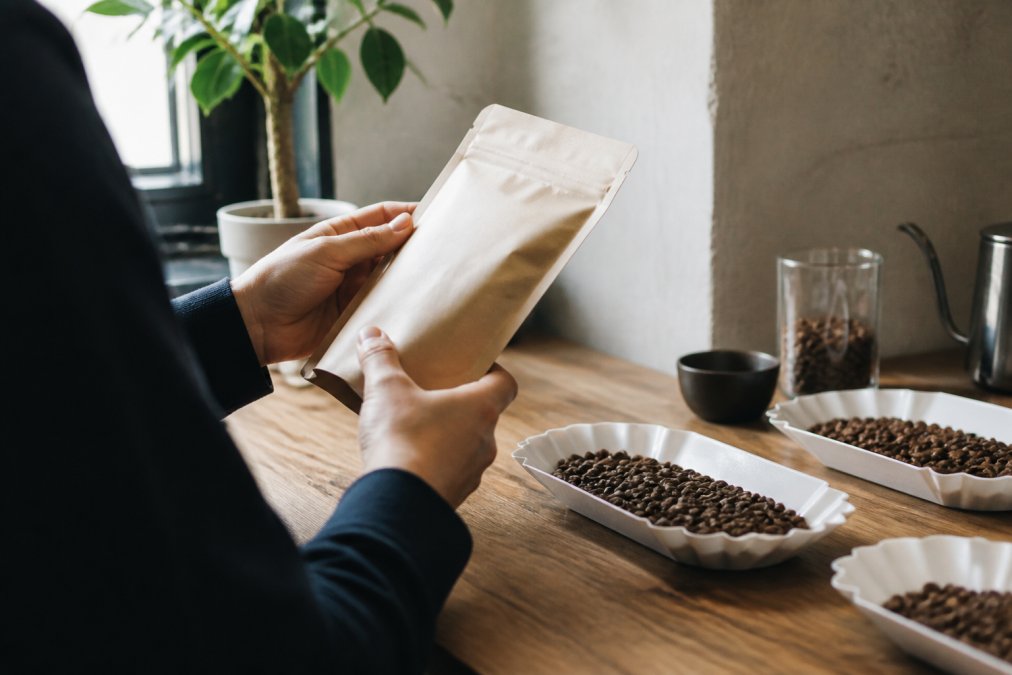 A coffee enthusiast's hands holding and examining a single-origin specialty coffee bag, with multiple roasted coffee beans visible on a white ceramic cupping plate in front of them, natural window lighting
