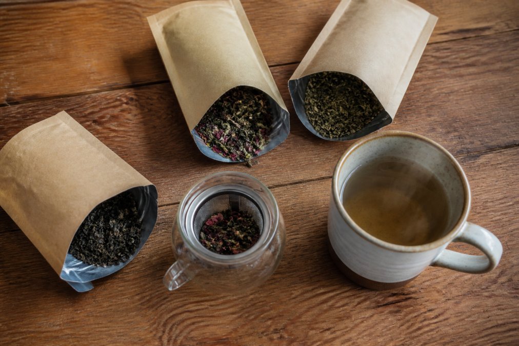 Overhead shot of three open kraft resealable tea pouches on a wooden table with loose leaf tea, a small glass infuser, and a steaming mug beside them