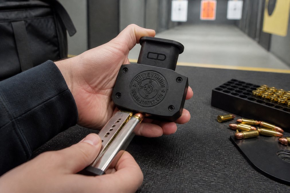 Close-up of hands inserting a 9mm magazine into a speed loader at an indoor shooting range, with rounds visible being fed into the loader opening, natural fluorescent lighting