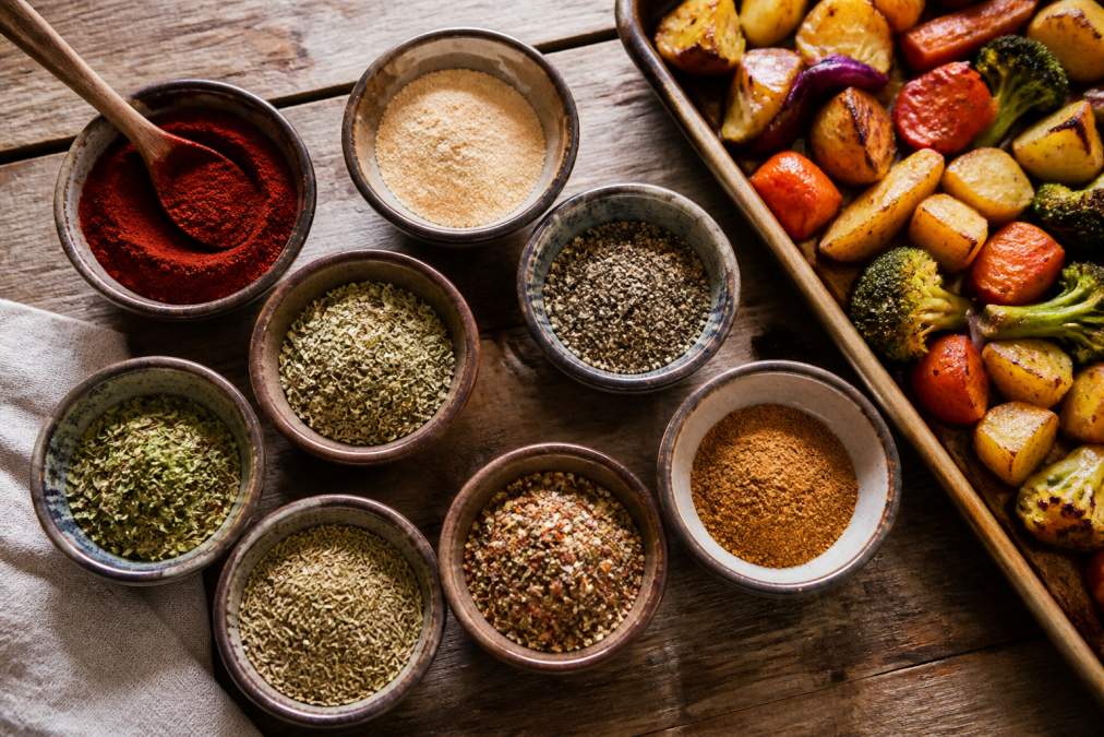 Overhead flat-lay of several small ceramic bowls filled with colorful spice blends including deep red smoked paprika, pale onion powder, cracked black pepper, and dried green herbs, arranged on a weathered wooden surface with a sheet pan of golden roasted vegetables partially visible at the edge, warm natural light