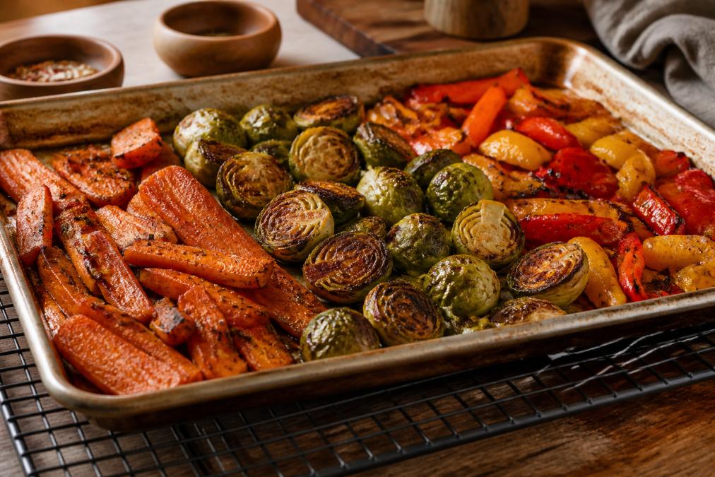 A sheet pan of golden-brown roasted vegetables including caramelized carrots, charred Brussels sprouts, and blistered bell peppers fresh from the oven, resting on a wire rack with visible crispy edges and deep color, warm kitchen lighting from overhead