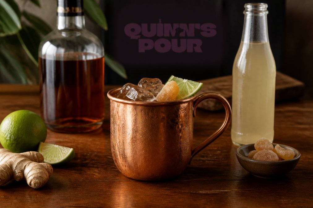 Freshly made Dark and Stormy cocktail in a copper mug, garnished with a lime wedge and candied ginger, with a bottle of spiced rum and ginger beer visible on a wooden bar surface
