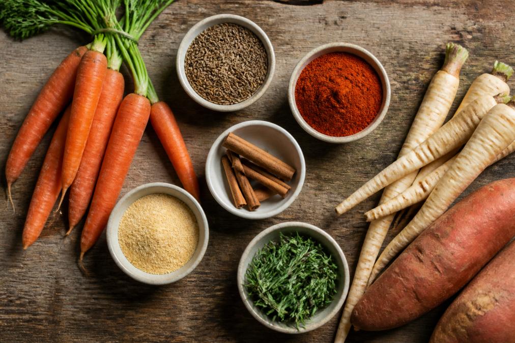 Overhead shot of a rustic wooden table with small bowls of cumin, smoked paprika, cinnamon sticks, garlic powder, and fresh rosemary, surrounded by raw carrots, parsnips, and sweet potatoes
