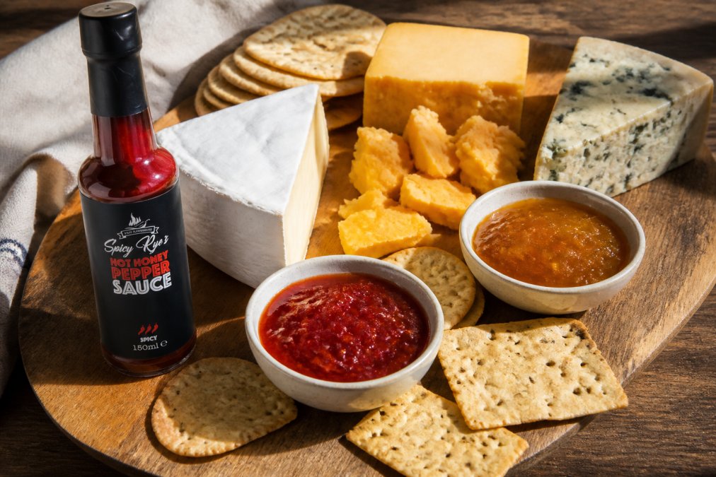 Close-up of a wooden cheese board with creamy white cheese, crumbly golden cheddar, and deep blue-veined cheese arranged beside small bowls of vibrant red and golden spicy jam, fresh crackers scattered across the board, natural afternoon light casting shadows