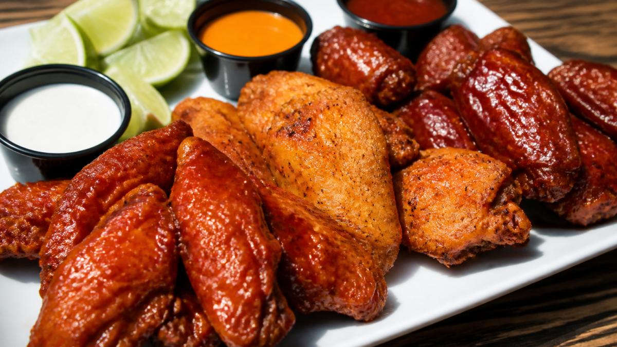 Close-up of crispy golden chicken wings coated in vibrant red and orange spicy sauces, arranged on a white plate with small bowls of dipping sauces and fresh lime wedges in natural daylight