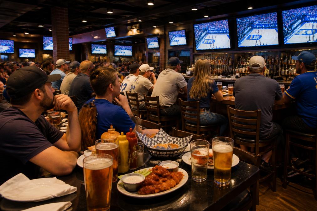 Interior of a sports bar with multiple large high-definition screens mounted on walls and positioned above the bar counter, customers seated at tables and bar stools with cold beers and plates of wings, energetic crowd watching a game with focused attention