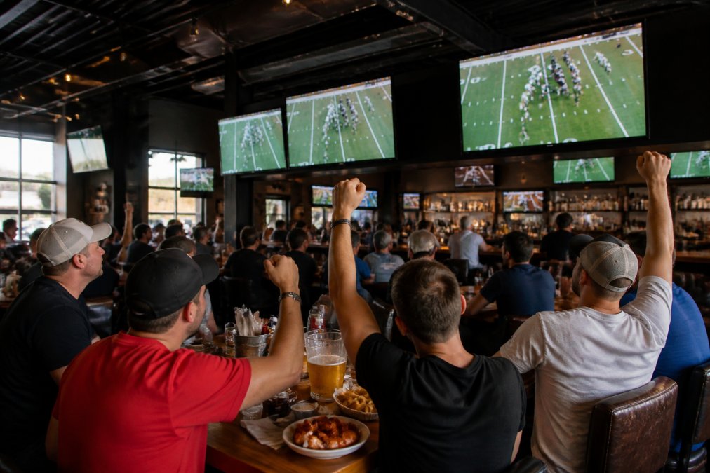 Wide shot of a lively sports bar interior with multiple large HD screens showing a football game, patrons cheering at tables with wings and beer pitchers under warm ambient lighting