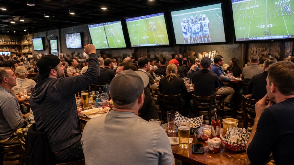 Lively sports bar interior with multiple large flat-screen TVs mounted on walls displaying live football games, patrons seated at bar stools and tables with cold beers, energetic crowd focused on the action with warm overhead lighting