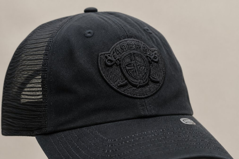 Close-up of vintage-style trucker cap showing embroidered team patch detail, mesh back panels, and worn-in fabric texture against neutral background