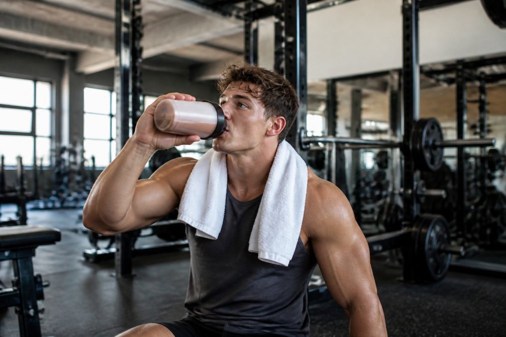 Athletic student drinking a protein shake immediately after finishing intense training session in the gym, towel around neck, natural gym lighting