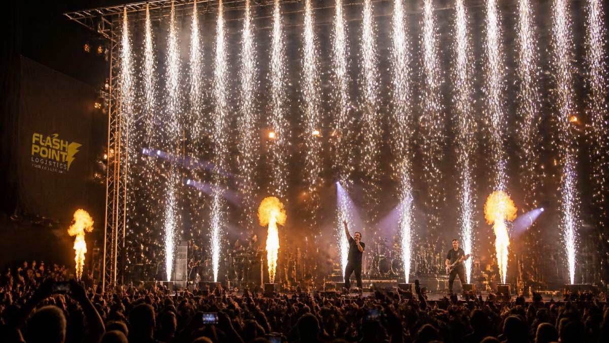 Professional concert stage featuring cold spark effects creating bright white cascading sparks above performers during high-energy moment with dark venue background and audience visible