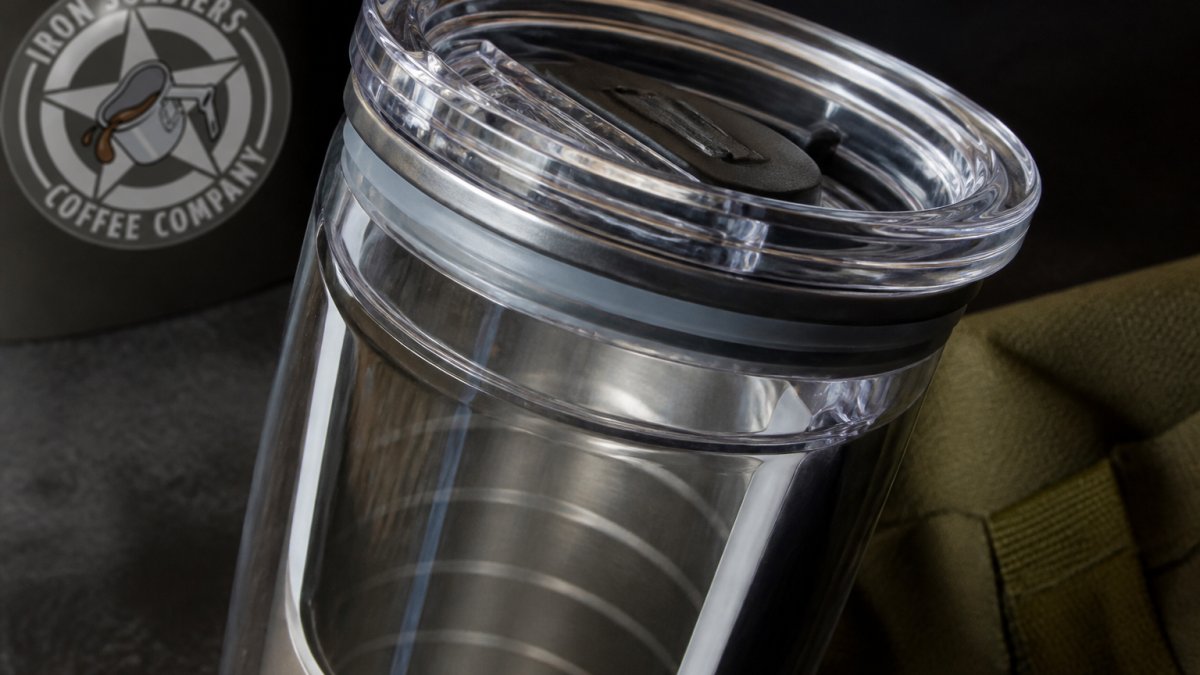 Close-up of a stainless steel tumbler with clear acrylic snap-on lid, showing the gasket seal mechanism and the interior plastic insulation visible through the transparent walls, positioned at a slight angle on a dark surface