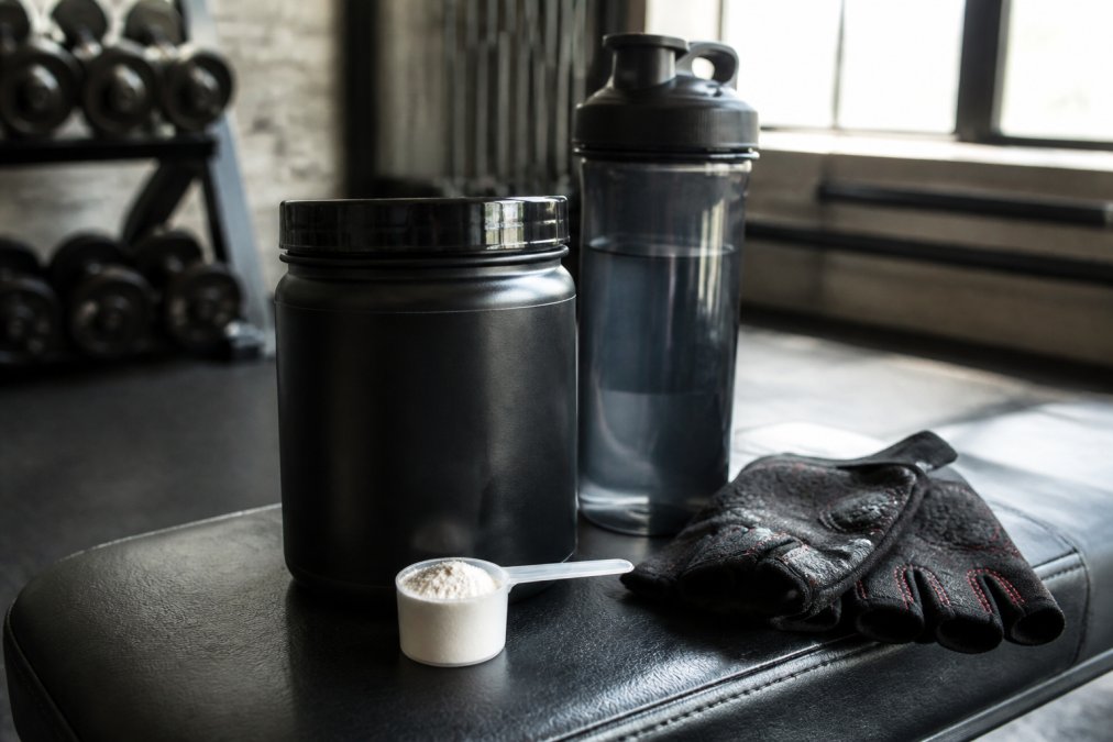 Close-up of a pre-workout supplement tub and scoop on a gym bench, with a filled water bottle and a pair of workout gloves placed beside it, natural gym lighting filtering through a window