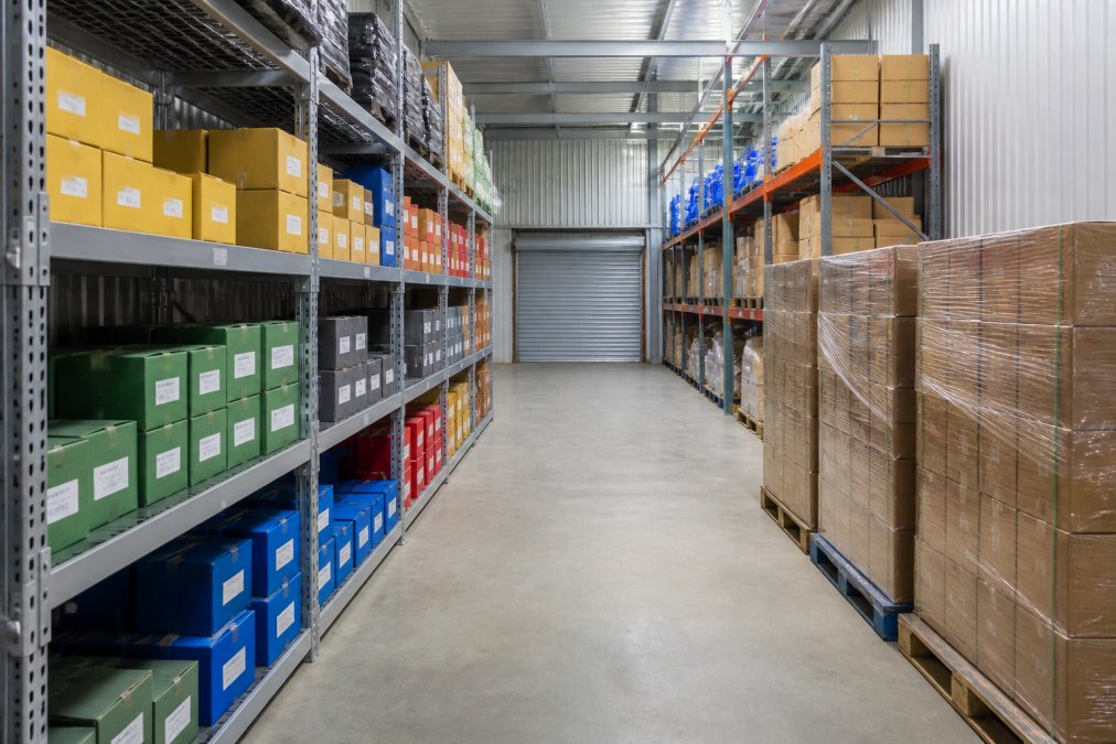 Interior view of a well-organised business storage unit showing colour-coded cardboard boxes arranged on metal shelving, clear labelled categories visible on each shelf, wide pathways between storage areas, and pallets of items stacked efficiently with proper weight distribution