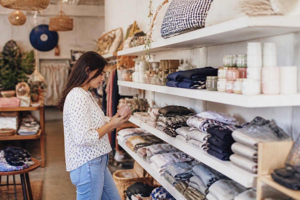 A neat display of new clothing, accessories, and home goods on bright shelves in a welcoming shop, with a person browsing the selection under warm lighting