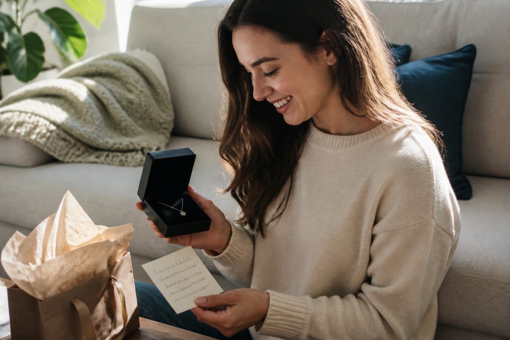Woman opening a jewelry gift box and admiring a personalized necklace with a heartfelt message card visible beside it, natural indoor lighting