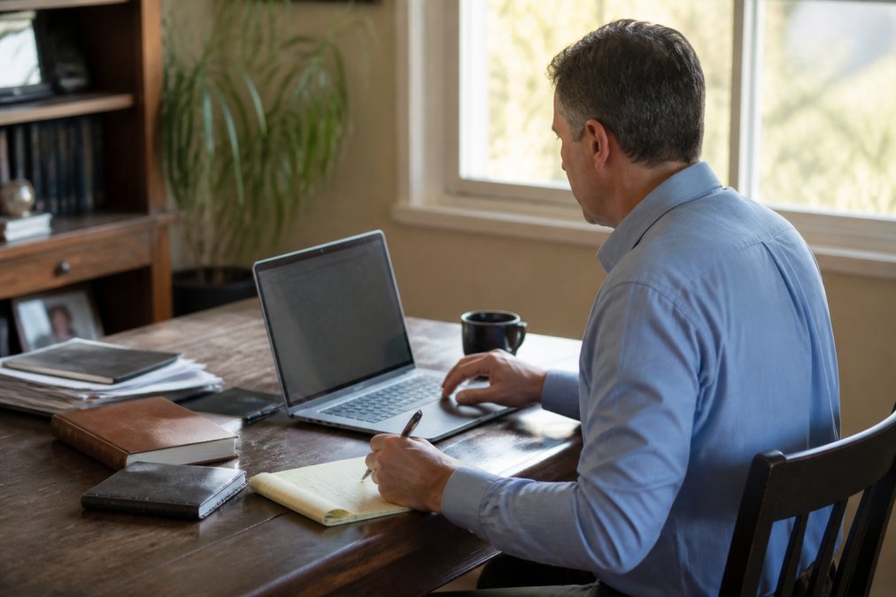 A person sitting at a desk with a laptop and notepad, researching law firms online in a home office, warm afternoon light through a window
