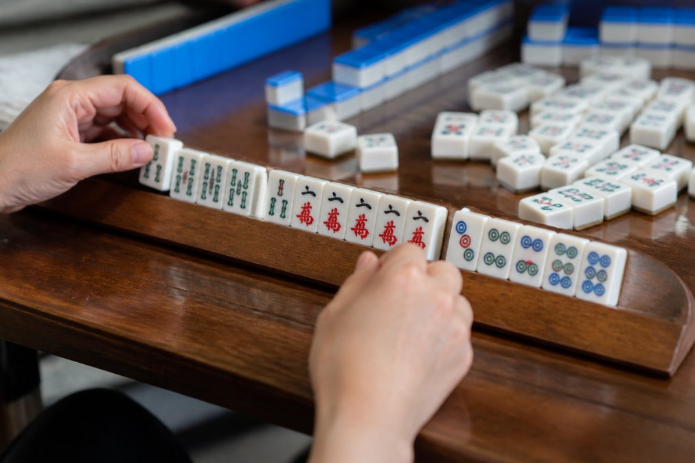 Close-up of hands arranging mahjong tiles on a wooden rack, showing organized groupings of tiles by suit and sequence with clear separation between bamboo, character, and dot suits during active gameplay