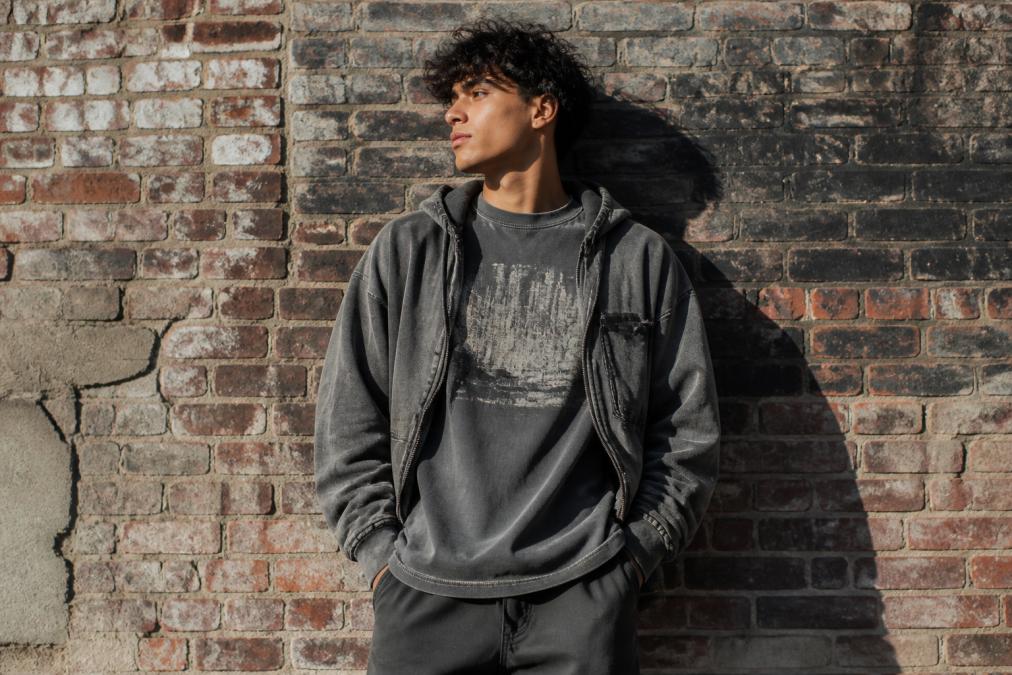 Young person standing confidently against industrial brick wall wearing oversized boxy tee with raw seams and city graphic, snow-washed hoodie layered over it, natural afternoon sunlight casting shadows on weathered brick backdrop