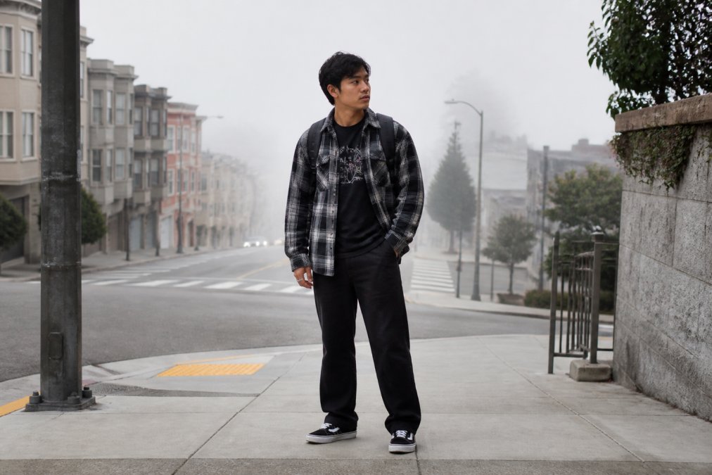 A young adult wearing a black graphic tee layered under an open flannel, standing on a foggy city street in the morning, with a backpack and casual sneakers, looking confident and relaxed