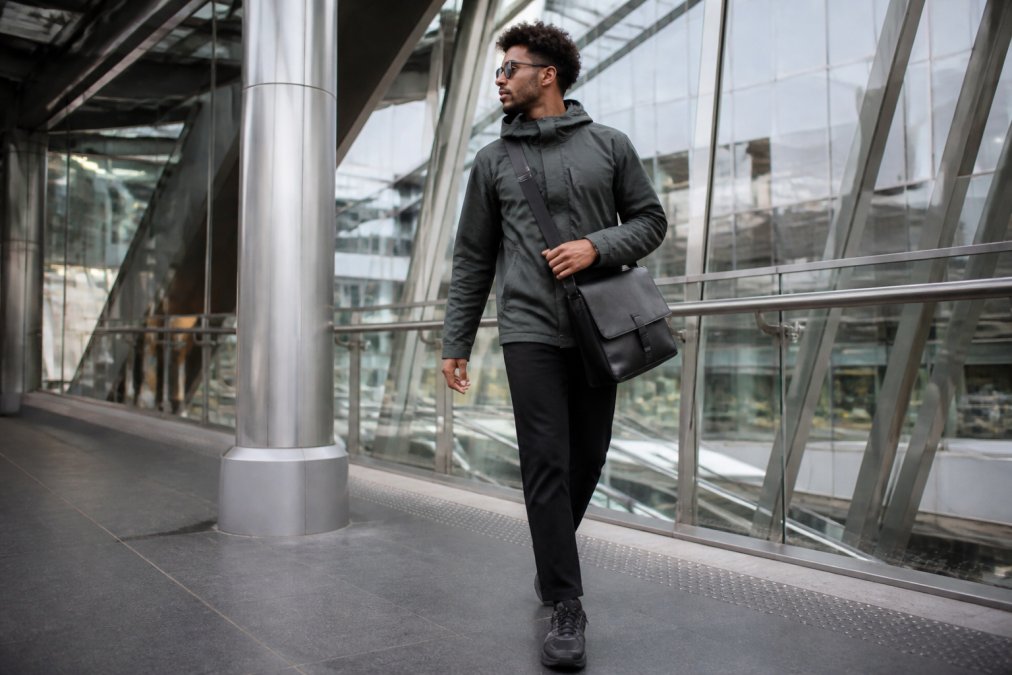 Professional commuter wearing layered technical streetwear walking through modern urban transit station with glass and steel architecture, carrying a structured messenger bag, wearing water-resistant jacket