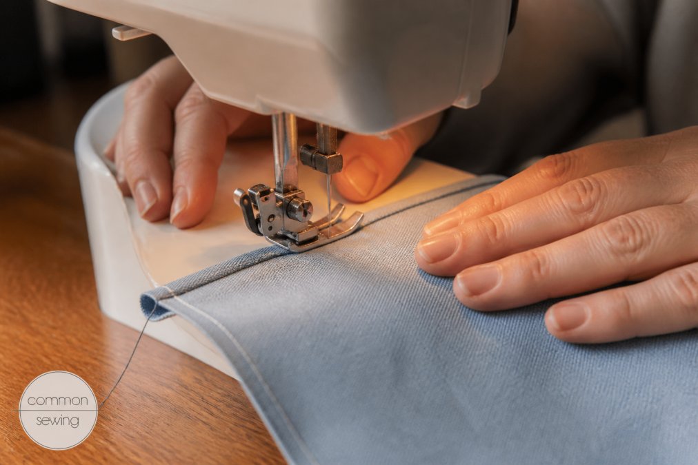 Close-up of a beginner's hands guiding fabric under a sewing machine presser foot, with thread and a partially sewn seam visible on a wooden table