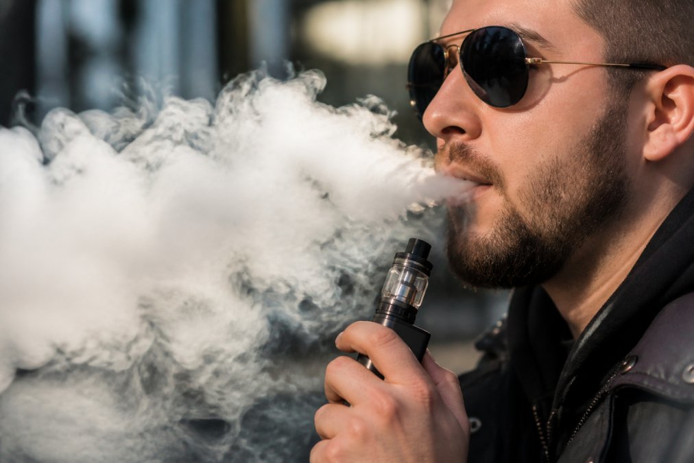 Person exhaling thick, visible vapor clouds from a high-wattage vape device, with the tank and coil clearly visible against a well-lit background