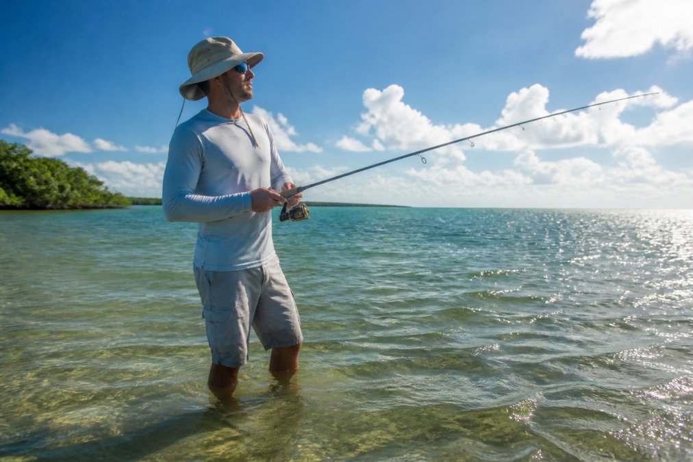 An angler wearing a long-sleeve moisture-wicking sun-protective shirt and wide-brim hat, standing in shallow water on a bright summer day, fishing rod in hand, sunlight reflecting off the water surface
