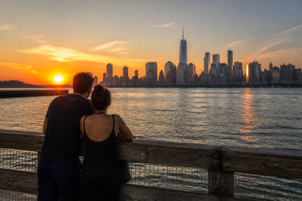 A couple leaning on a weathered wooden railing at a waterfront pier, Manhattan skyline glowing orange behind them as the sun sets over open water